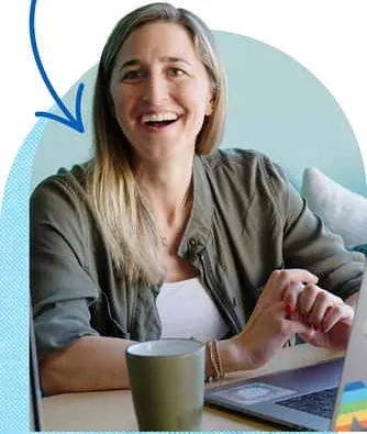 Laughing blonde woman in olive jacket works at laptop with mug