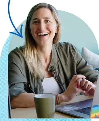 Laughing blonde woman in olive jacket works at laptop with mug