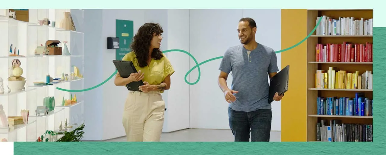 Two coworkers carrying laptops walk and chat through a colorful office.