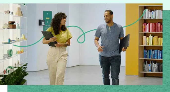 Two coworkers carrying laptops walk and chat through a colorful office.
