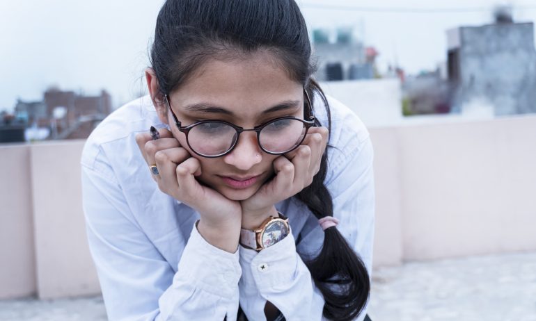 A female student, wearing round black-frame glasses and a white shirt, rests her chin in her hands