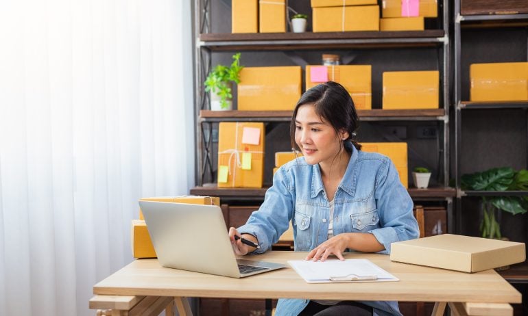 Young woman with her dark hair pulled back works at a laptop with paperwork on her desk.