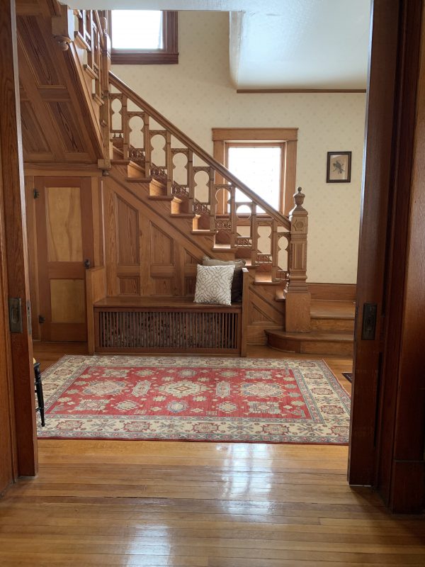 A oak staircase to the second floor, a red and tan patterned rug and shiny hardwood floors are shown in the Victorian-style house.