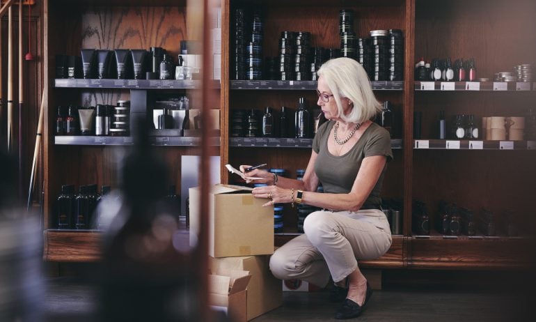Senior saleswoman holding paper while crouching by boxes in deli