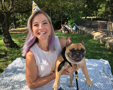 Chelsea Amengual smiles and wears a party hat while sitting outside on a blanket and holding her French bulldog. 