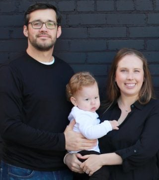 CJ Mantilla stands in front of a dark brick backdrop in dark-rimmed glasses. His right hand rests on the back of his baby son, whom Steffa Mantilla holds on one hip. She smiles at the camera as the baby grips her collar.
