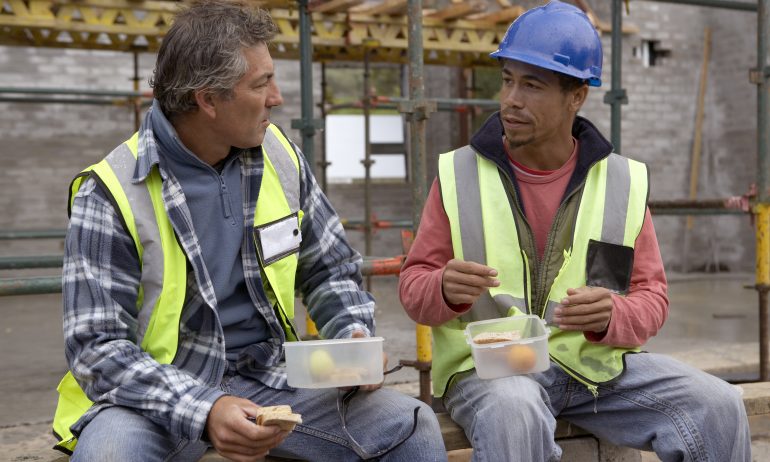 Two construction workers sitting down on a residential building site while eating their lunch.