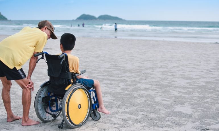 Rear View Of Man By Disabled Boy On Wheelchair At Beach