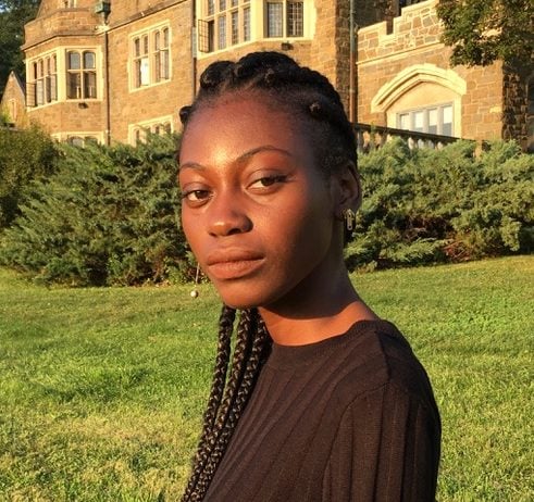 Funto stands in profile, looking across her left shoulder into the camera, outside on a sunny day.