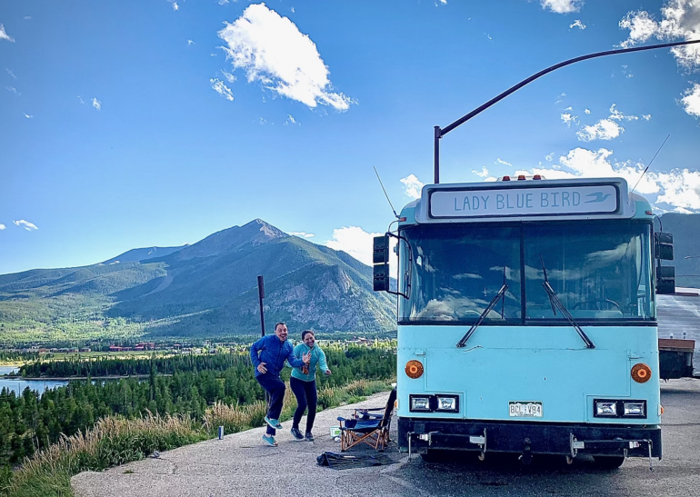Steve and Carly Schneiter make running motions outside their blue bus that's parked near a lake. 
