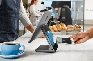 A customer uses her phone to checkout with a digital wallet. She holds her cell phone against the customer-facing screen of the register while the cashier stands behind the counter of a bakery.