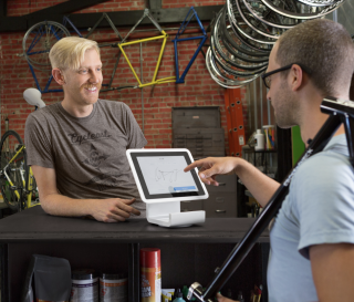 A customer signs on a Square Stand POS system screen while a merchant smiles at him from behind the counter.