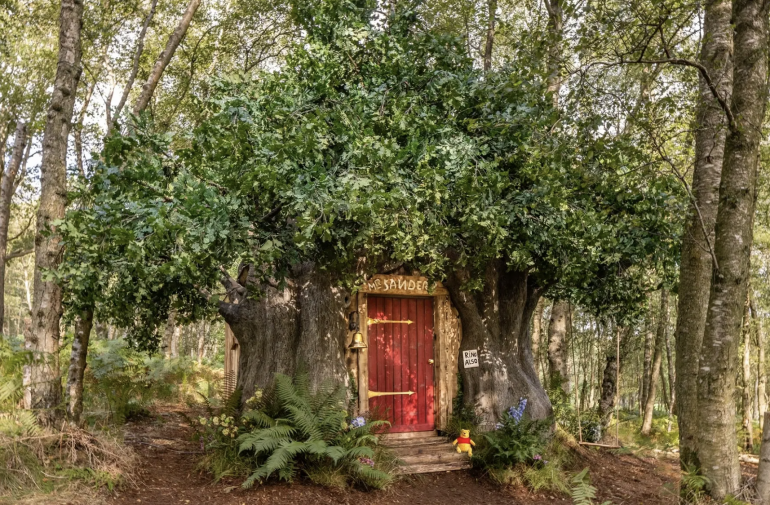 A small house resembling a tree sits in the woods with a red door and small Winnie the Pooh stuffed figure on the front steps. 