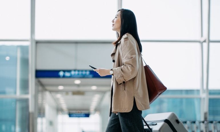 Young Asian woman carrying suitcase and holding smartphone on hand, walking in airport terminal. Ready to travel. Travel and vacation concept. Business person on business trip
