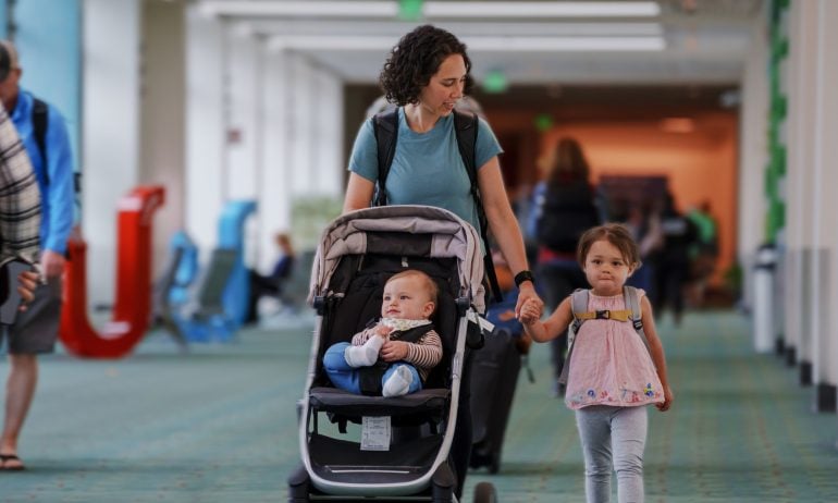Young Eurasian woman holds hands with her preschool aged daughter and pushes her 9 month old son in a stroller while walking through airport.