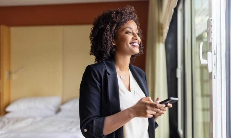 African businesswoman arrived at the hotel taking a rest from the trip. Female entrepreneur standing by hotel room window with her mobile phone.