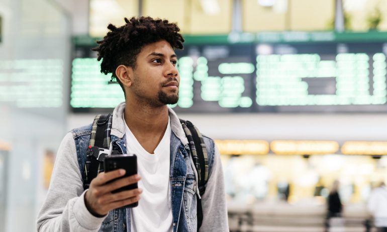 Young man inside the airport holding a mobile phone.young afro man holding a mobile phone looking at the departures of planes in an airport