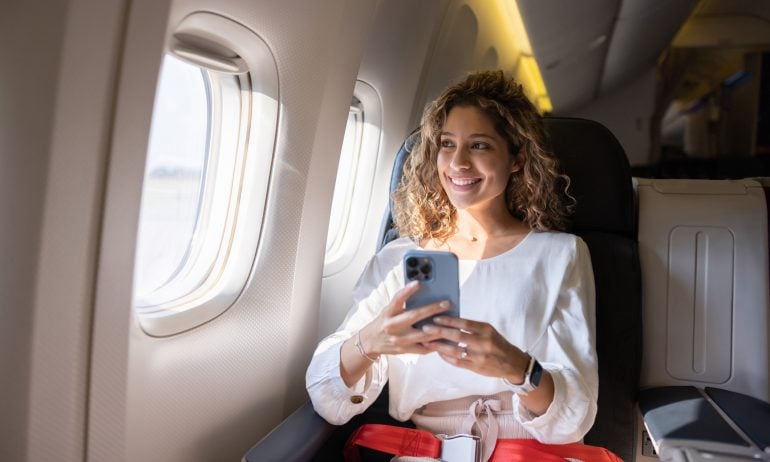 Latin American female traveler using her cell phone in an airplane and looking through the window