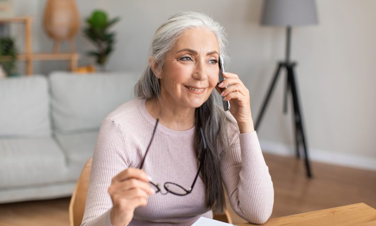 Smiling caucasian mature gray-haired lady hold glasses speaks by phone with client in living room interior