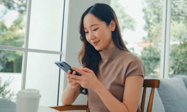 A young Asian woman is sitting in a cafe, using her smartphone to bank online.