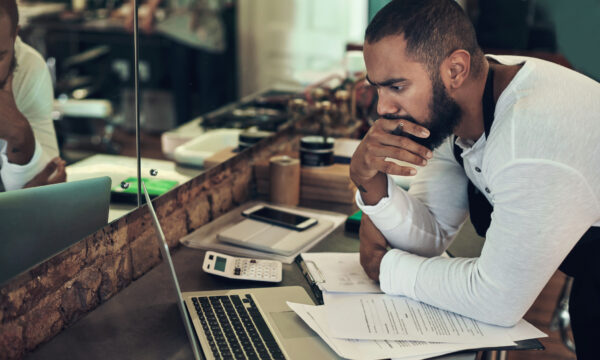 Young barber looking contemplative while using his laptop to calculate his finances in his salon