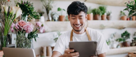 Asian male flower shop owner browsing business bank accounts on his tablet.