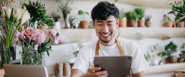 Asian male flower shop owner browsing business bank accounts on his tablet.