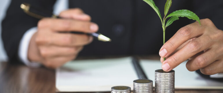 Hand on desk with growing piles of money thanks to smart investing.