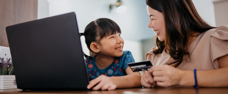 Young Asian mother and daughter manage a kids bank account on laptop.