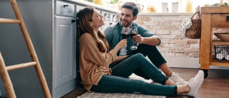 Young couple with wine glasses sits on kitchen floor discussing how to invest in stocks.