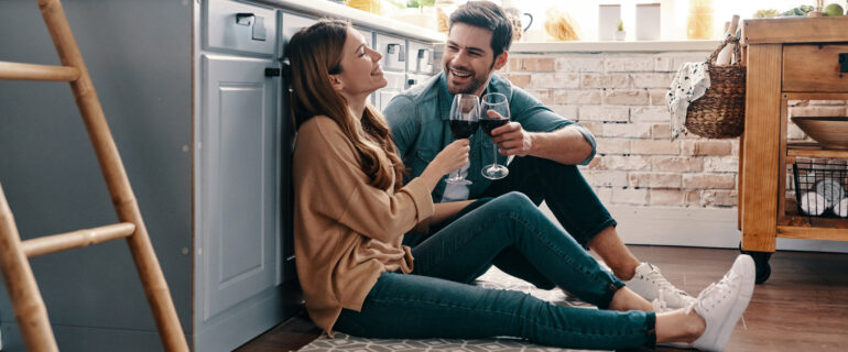 Young couple with wine glasses sits on kitchen floor discussing how to invest in stocks.