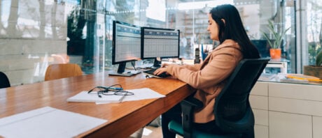 Investment banker sits at desk looking at monitors with data on them.