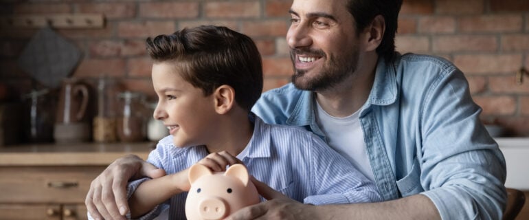 Happy dad and little tween boy sit at the table hug hold piggy bank look at the distance dream of their growing term deposits.