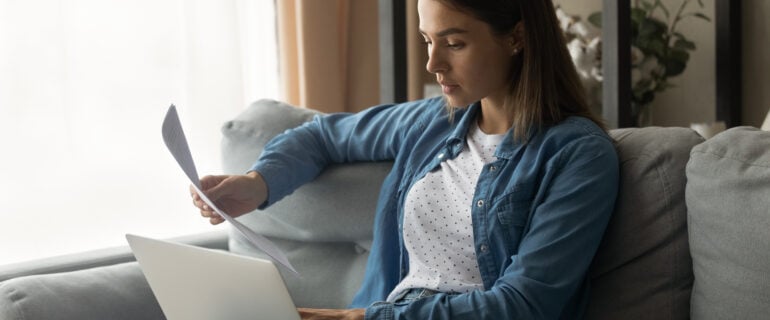 A young millennial woman reads the paper hardcopy of the personal loan agreement and checks information online using a laptop.