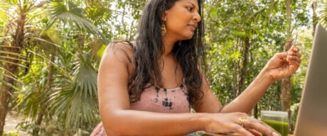 A multiracial woman in a tropical garden using her American Express Green Card Review to make a booking on her laptop.