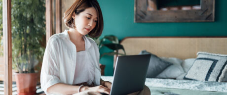 A young Asian woman reading the RBC TFSA review on her laptop while sitting on the bed.