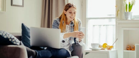 A millennial girl checks her Scotiabank gic performance on her laptop, lounging on her sofa and texts her friends about it.