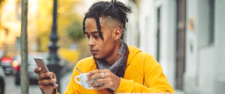 An international student checking his bank application status on his smartphone while having coffee and studying at a coffee shop.