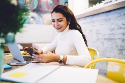 A happy South Asian female student is texting someone about her Motive Financial GIC performance that she checked on her laptop.