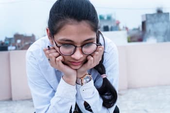 A female student, wearing round black-frame glasses and a white shirt, rests her chin in her hands
