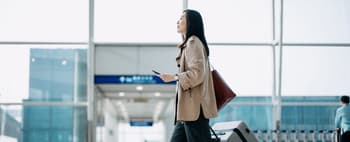 Young Asian woman carrying suitcase and holding smartphone on hand, walking in airport terminal. Ready to travel. Travel and vacation concept. Business person on business trip