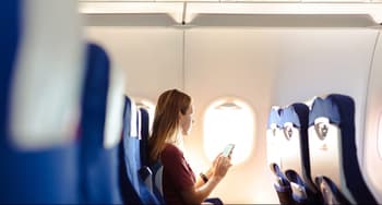 Young woman sitting on the aircraft seat near the window during the flight in the airplane. She is using smart phone with a blank screen