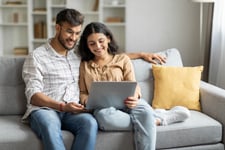 A loving, happy Indian couple sitting on the couch at home, embracing, using laptop computer.