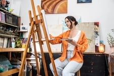 Mid-adult woman painting on a canvas while holding an artist's pallet at home.