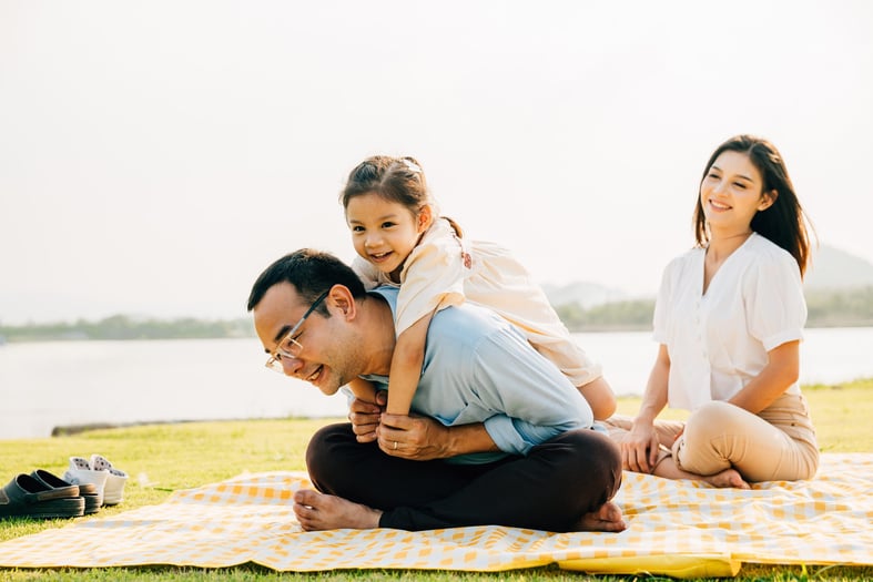 Happy Asian family sitting on a picnic blanket in the park. Little girl riding father back enjoying outdoors together.