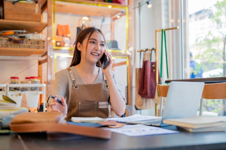A female leather bag designer is talking on her mobile while checking her business loan balance on her laptop.