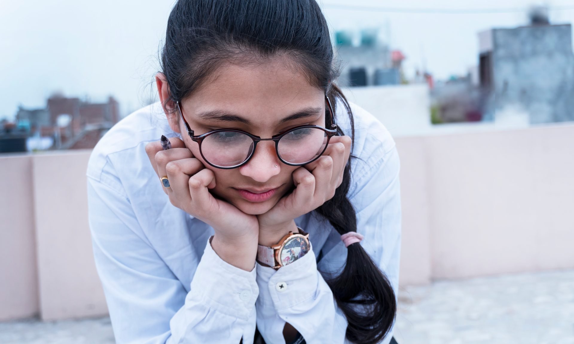A female student, wearing round black-frame glasses and a white shirt, rests her chin in her hands