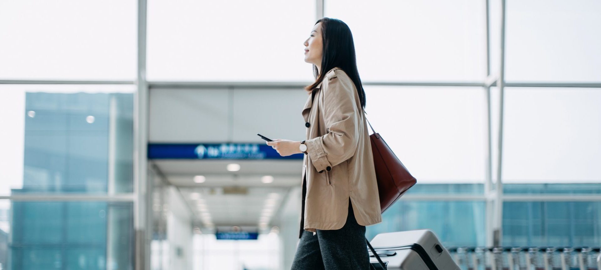 Young Asian woman carrying suitcase and holding smartphone on hand, walking in airport terminal. Ready to travel. Travel and vacation concept. Business person on business trip