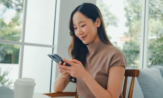 A young Asian woman is sitting in a cafe, using her smartphone to bank online.