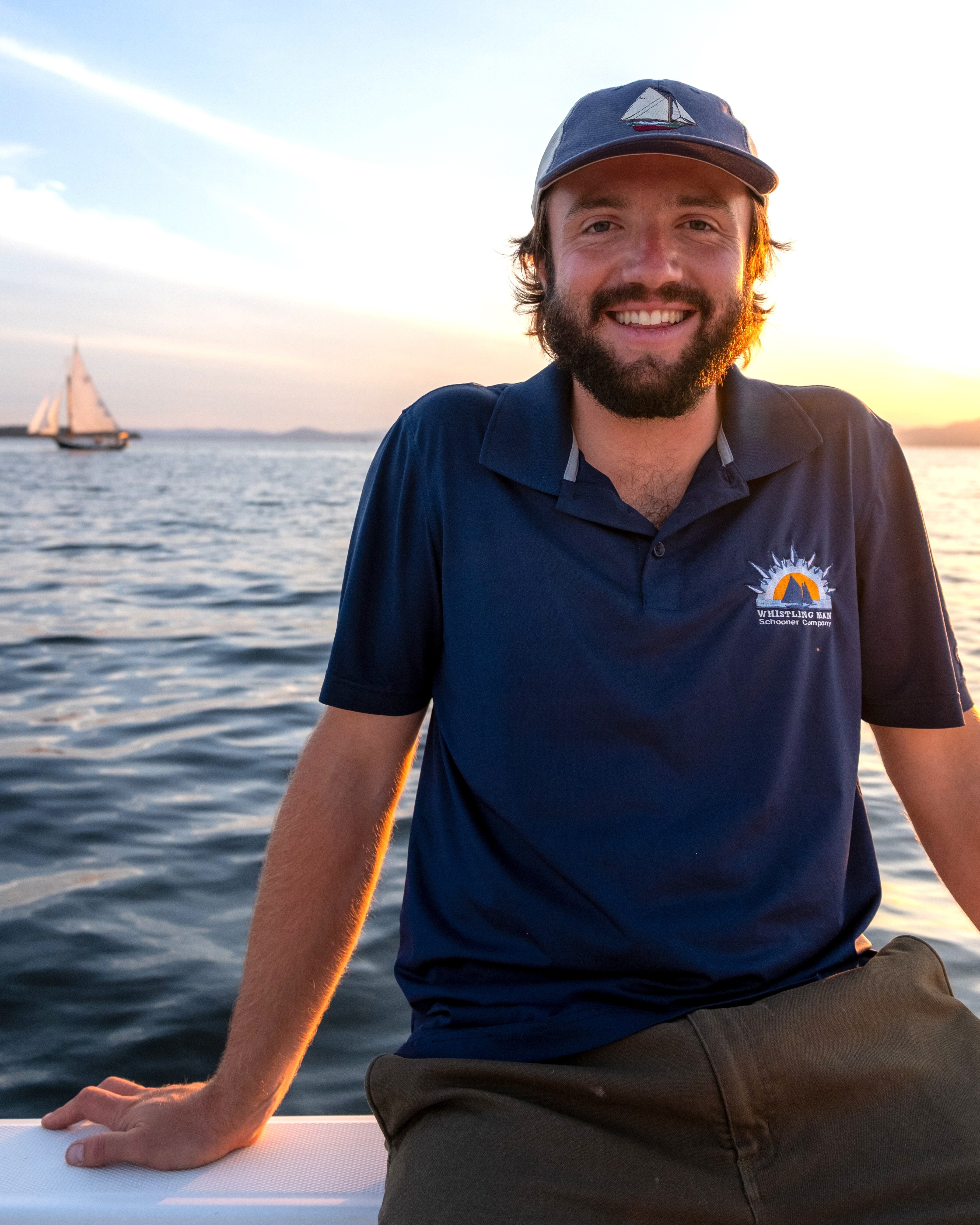 Bearded business owner wearing baseball cap sits on sailboat smiling at camera while sun sets behind him